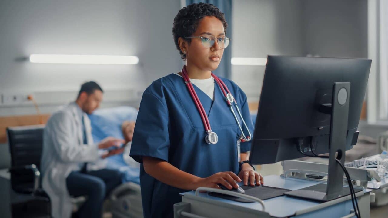 Hospital Ward: Professional Smiling Black Female Head Nurse or Doctor Wearing Stethoscope Uses Medical Computer. In the Background Patients in Beds Recovering Successfully after Sickness and Surgery